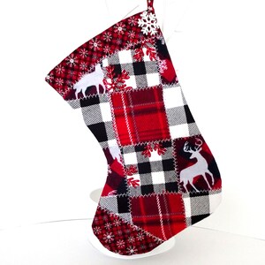 May include: A Christmas stocking with a patchwork design featuring red plaid, black and white checkered patterns, and white deer silhouettes. The stocking has a red snowflake pattern at the top and a white loop for hanging.