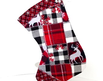 Pode incluir: Uma meia de Natal com um design de patchwork com xadrez vermelho, padrões de xadrez preto e branco e silhuetas de veados brancos. A meia tem um padrão de flocos de neve vermelhos na parte superior e uma argola branca para pendurar.