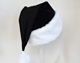 Könnte beinhalten: Eine schwarze Weihnachtsmütze mit einem breiten, flauschigen weißen Kunstfellbesatz am Rand und einem passenden weißen Bommel an der Spitze. Der Hauptteil der Mütze besteht aus einem einfarbigen schwarzen Stoff, der einen Kontrast bildet.
