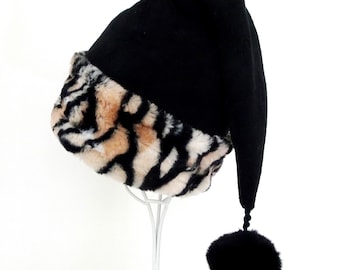 May include: A black pointed hat featuring a wide, plush faux fur band with a black, tan, and white tiger-like animal print pattern. The hat has a long, drooping crown that terminates in a solid black pom-pom attached by a braided cord.