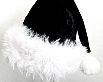 Könnte beinhalten: Eine schwarze Samtmütze mit einem breiten, flauschigen weißen Kunstfellbesatz am Rand und einem passenden weißen Bommel an der Spitze. Die Mütze hat eine klassische konische Form mit einer weichen, strukturierten Oberfläche des schwarzen Stoffes.