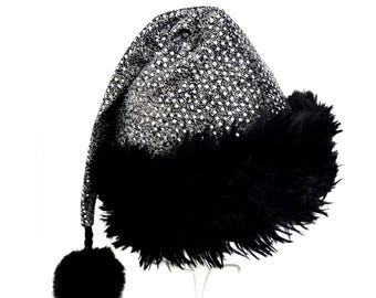 May include: A conical hat featuring a shimmering silver and black textured fabric with a dense, fluffy black faux fur trim around the base. The hat tapers to a point and includes a dangling black pom-pom attached to the tip by a thin cord.