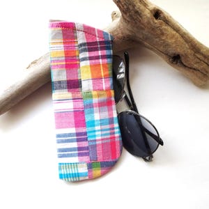 K&ouml;nnte beinhalten: Ein buntes Sonnenbrillenetui aus Patchwork-Stoff in Rosa, Blau, Gelb und Orange. Das Etui enth&auml;lt eine schwarze Sonnenbrille. Im Hintergrund ist ein St&uuml;ck Treibholz zu sehen.