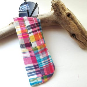 K&ouml;nnte beinhalten: Ein bunt kariertes Sonnenbrillenetui in Rosa, Blau, Gelb und Orange. Das Etui besteht aus Patchwork-Stoff und enth&auml;lt eine Sonnenbrille mit schwarzem Gestell und verspiegelten Gl&auml;sern. Im Hintergrund liegt ein St&uuml;ck Treibholz.