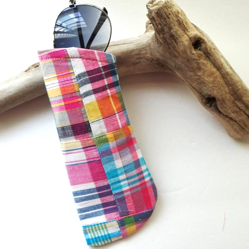 K&ouml;nnte beinhalten: Ein bunt kariertes Sonnenbrillenetui in Rosa, Blau, Gelb und Orange. Das Etui besteht aus Patchwork-Stoff und enth&auml;lt eine Sonnenbrille mit schwarzem Gestell und verspiegelten Gl&auml;sern. Im Hintergrund liegt ein St&uuml;ck Treibholz.