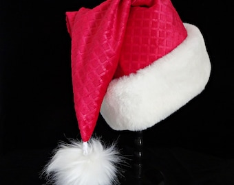 May include: A festive Santa hat with a vibrant red, textured fabric and a plush white trim. The hat features a fluffy white pom-pom at the end. The hat is displayed on a black stand against a black background.