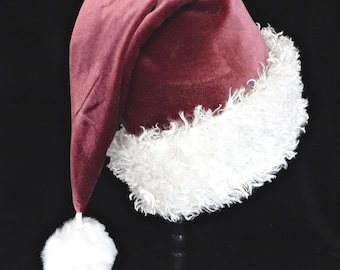 Può includere: Un cappello da Babbo Natale in velluto bordeaux con finiture bianche e soffici e un pompon abbinato. Il cappello è esposto su un supporto nero, mettendo in risalto il suo design tradizionale e il suo fascino festivo.