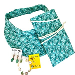 Puede incluir: Una visera de sol floral de color turquesa, una bolsa a juego de color turquesa floral con cordón blanco, un par de pendientes de lágrima verde y una pulsera de cuentas verdes y blancas con un anillo de plata. Las joyas están etiquetadas con "de la Mar" y la pulsera con "JelaMar Sea Ring".