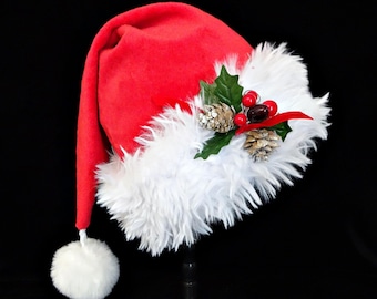 Puede incluir: Un gorro de Papá Noel rojo con un ribete blanco y esponjoso y un pompón blanco. El gorro está decorado con una ramita de acebo, bayas rojas y dos pequeñas piñas. El gorro se muestra sobre un fondo negro.