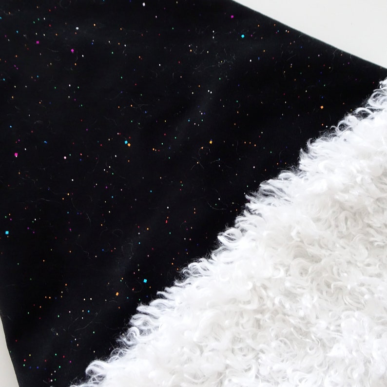 May include: Black fabric with a constellation of colorful glitter dots, paired with a fluffy white faux fur trim. The contrasting textures and colors create a visually appealing design. The fabric has a soft, slightly textured appearance.
