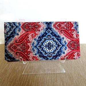 May include: A rectangular wallet with a vibrant paisley pattern in red, white, and navy blue. The wallet is displayed on a clear acrylic stand, showcasing its intricate design and colour scheme. The fabric features a detailed, symmetrical design.