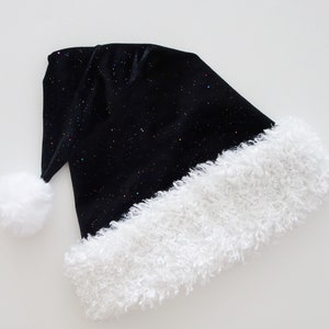 May include: A black Santa hat with a white, fluffy trim and a matching pom-pom. The hat is covered in tiny, colorful glitter, giving it a starry appearance. The hat is made of a soft, velvet-like material.