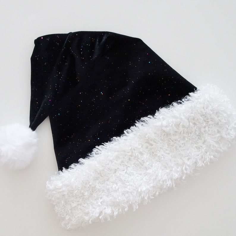 May include: A black Santa hat with a white, fluffy trim and a matching pom-pom. The hat is covered in tiny, colorful glitter, giving it a starry appearance. The hat is made of a soft, velvet-like material.
