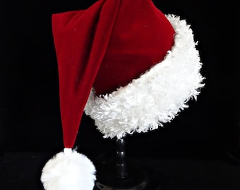 May include: A deep red velvet Santa hat featuring a thick, curly white faux fur trim around the base and a large, fluffy white pom-pom attached to the pointed tip. The hat is displayed on a black stand against a solid black background.