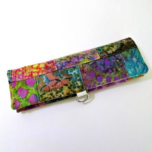 May include: A colorful, patchwork-style clutch with a white strap and silver ring. The fabric features vibrant hues of pink, green, purple, and blue in a batik-like pattern. The rectangular clutch is closed and appears to be made of fabric.