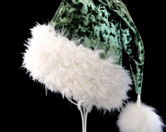 May include: A conical hat made of dark green crushed velvet fabric. The base of the hat features a wide, thick band of long, shaggy white faux fur. A matching white faux fur pom-pom is attached to the pointed tip of the hat by a short cord.