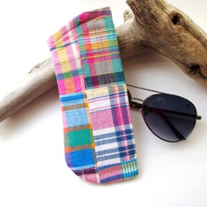May include: A colorful patchwork sunglasses case in pink, blue, yellow, and green plaid fabric. The case is open, revealing a pair of aviator sunglasses with dark lenses and a silver frame. The case is next to a piece of driftwood.