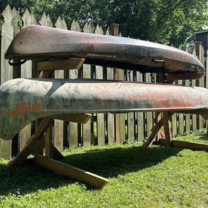 May include: Two canoes are stacked on top of each other on a wooden rack. The canoes are both in a worn condition, with one being a dark brown color and the other being a camouflage pattern with green, brown, and orange colors. The rack is made of wood and has a simple design.