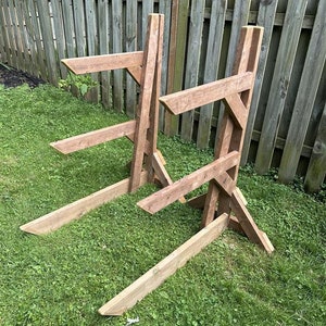 May include: Two wooden A-frame stands for storing long boards, such as surfboards or paddleboards. The stands are made of natural wood and are placed on a grassy lawn.