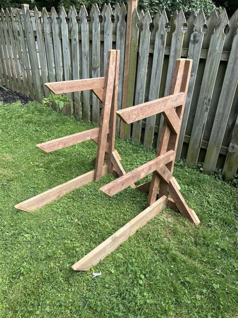 May include: Two wooden A-frame stands for storing long boards, such as surfboards or paddleboards. The stands are made of natural wood and are placed on a grassy lawn.