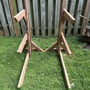 May include: Two wooden A-frame stands with horizontal supports, likely for holding firewood or other items. The stands are made of natural brown wood and are positioned on a grassy lawn.