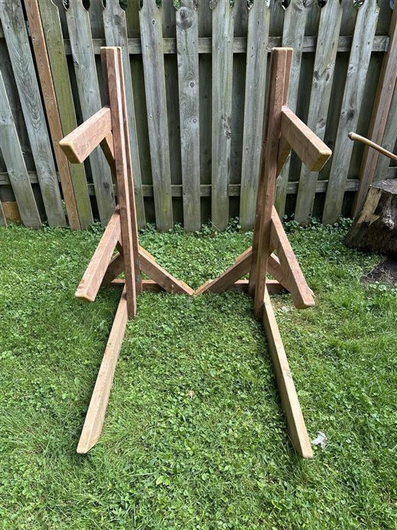 May include: Two wooden A-frame stands with horizontal supports, likely for holding firewood or other items. The stands are made of natural brown wood and are positioned on a grassy lawn.