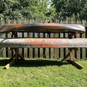 May include: Two canoes stacked on top of each other on a wooden stand. The canoes are both brown and orange with a faded paint job. The stand is made of wood and has a rustic look. The canoes are in a backyard setting with a wooden fence in the background.