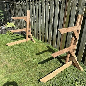 May include: Two wooden A-frame stands for storing firewood. The stands are made of brown wood and are placed on a green grassy lawn in front of a wooden fence.