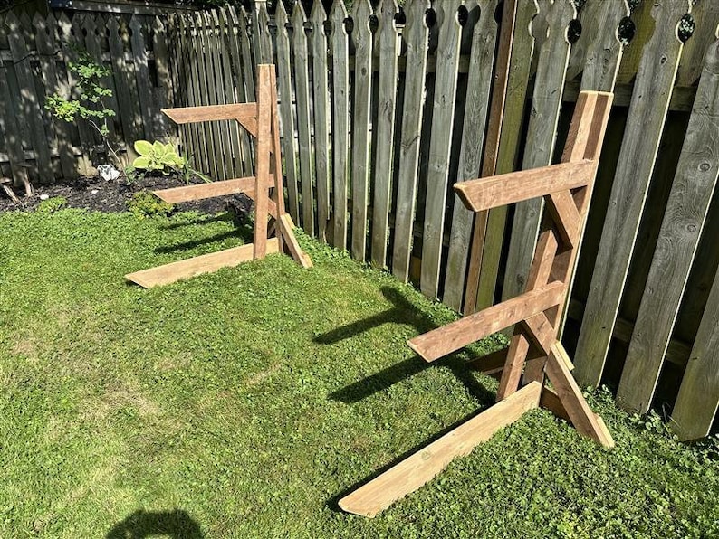 May include: Two wooden A-frame stands for storing firewood. The stands are made of brown wood and are placed on a green grassy lawn in front of a wooden fence.