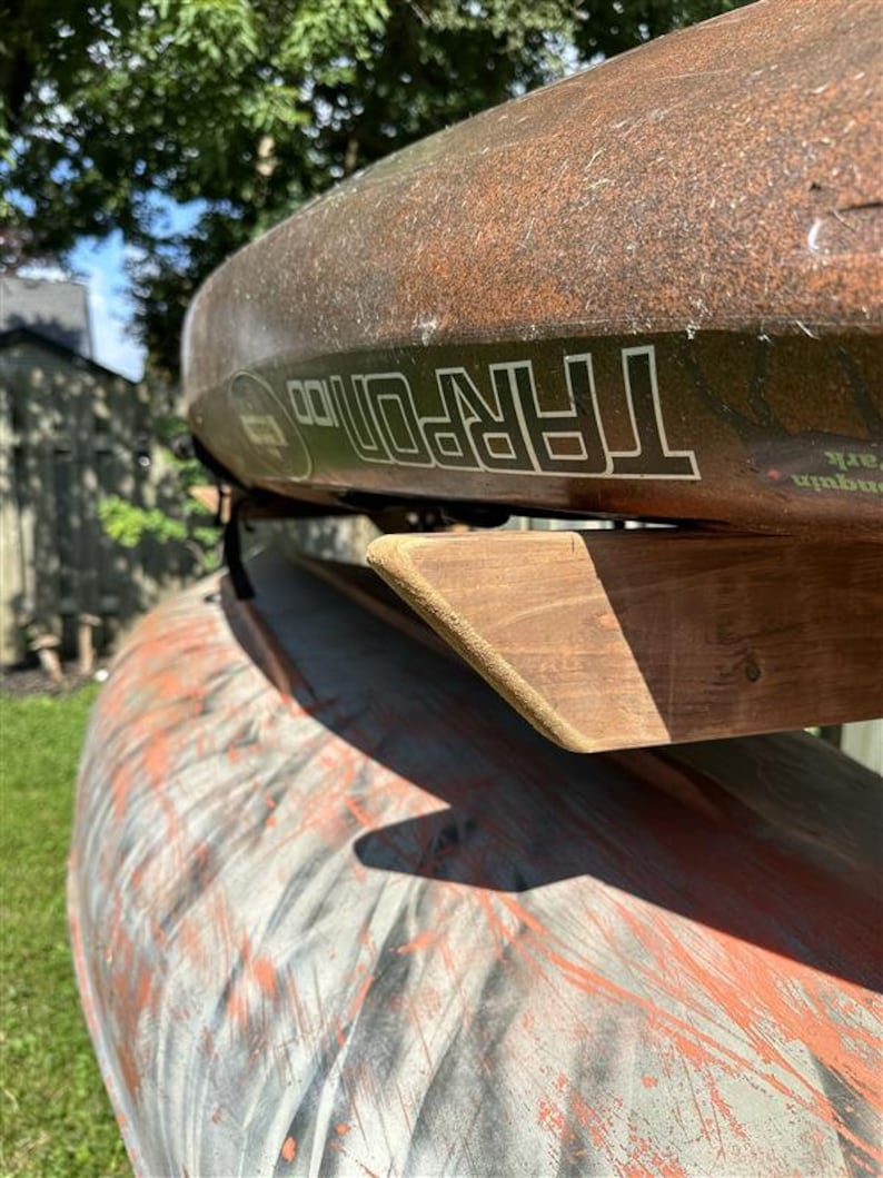 May include: Two brown kayaks stacked on a wooden rack. The top kayak has the word "TARPON" printed on the side.