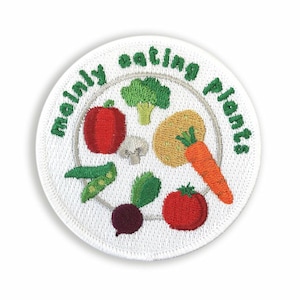 May include: White embroidered patch with the text "Mainly eating plants" in a circle around a plate with various vegetables, including tomatoes, broccoli, carrots, peas, and a beet.