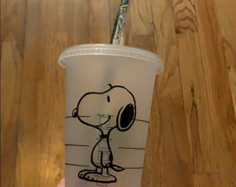 May include: A clear plastic Starbucks cold beverage cup with a Snoopy cartoon graphic. The cup has a clear lid and a green and white striped straw. The cup is sitting on a wooden surface.
