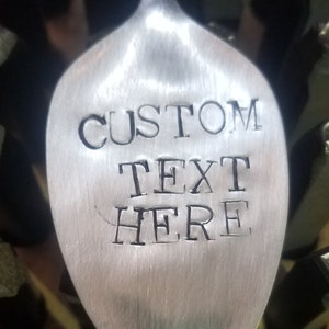 Puede incluir: Una cuchara de plata con las palabras "CUSTOM TEXT HERE" grabadas en ella. La cuchara está rodeada de sellos de letras metálicas.