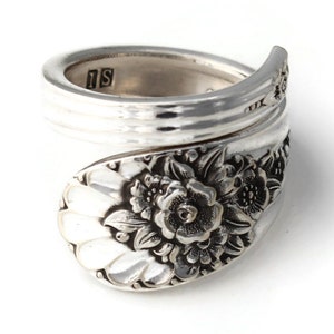 May include: A silver spoon ring with a floral design. The ring is made from a spoon and features a detailed flower and leaf pattern on the spoon's bowl. The ring is wrapped around the finger.