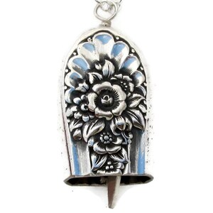 May include: A silver bell pendant featuring a floral design. The bell is teardrop shaped and has a detailed floral pattern.