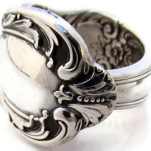 May include: A silver ring crafted from repurposed silverware. The ring features ornate floral and scrollwork details, with a polished finish. The design incorporates the spoon's bowl and handle, creating a unique, vintage-inspired accessory.