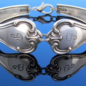 Könnte beinhalten: Versilbertes Löffelarmband mit aufwendigen Details. Das Armband besteht aus recyceltem Besteck und zeigt Löffel mit eingravierten Initialen. Das Design umfasst einen Karabinerverschluss und dekorative Verzierungen, die eine Vintage-Ästhetik widerspiegeln.