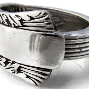 Puede incluir: Un anillo de plata hecho a partir de una cuchara reutilizada. El anillo tiene una banda ancha con un diseño decorativo en la parte superior e inferior. La banda es lisa y brillante.
