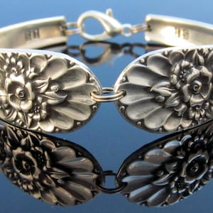 May include: A silver bracelet made from a vintage spoon with a floral design. The bracelet is connected with a gold-toned clasp.