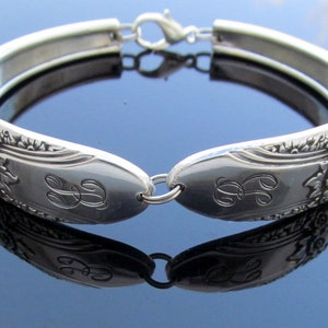 Könnte beinhalten: Ein silbernes Armband aus einer wiederverwendeten Löffel mit floralen Details und einem gravierten Monogramm. Das Armband ist offen und hat einen Verschluss.