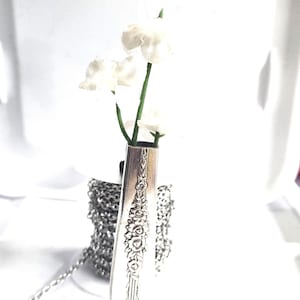 May include: A silver pendant necklace featuring a repurposed vintage fork, with a floral design. The fork holds small white flowers. The necklace chain is silver and visible in the image.