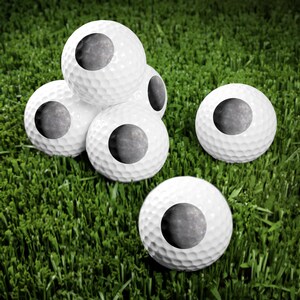 May include: A collection of five white golf balls, each featuring a circular graphic of the moon's surface. The golf balls have a dimpled texture and are arranged in a cluster, with one ball positioned separately in the foreground.