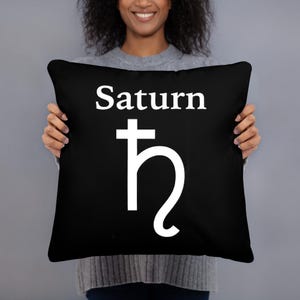 May include: A square black throw pillow featuring the white text Saturn centred at the top, positioned above the white astronomical symbol for the planet Saturn, which consists of a cross with a curved tail extending from the bottom.