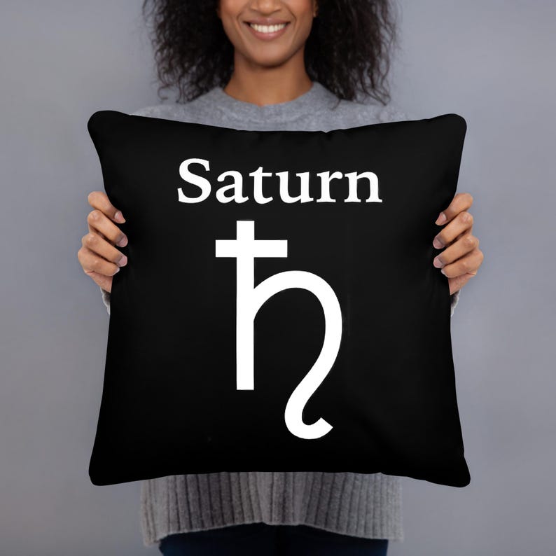 May include: A square black throw pillow featuring the white text Saturn centred at the top, positioned above the white astronomical symbol for the planet Saturn, which consists of a cross with a curved tail extending from the bottom.
