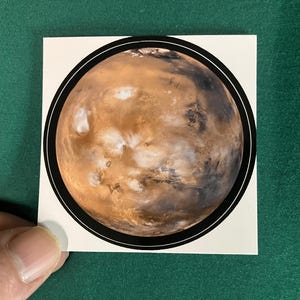 Puede incluir: Una pegatina cuadrada que presenta una ilustraci&oacute;n circular del planeta Marte. El planeta est&aacute; representado en tonos naranja &oacute;xido, marr&oacute;n y blanco, encerrado dentro de un grueso borde circular negro sobre un fondo blanco.