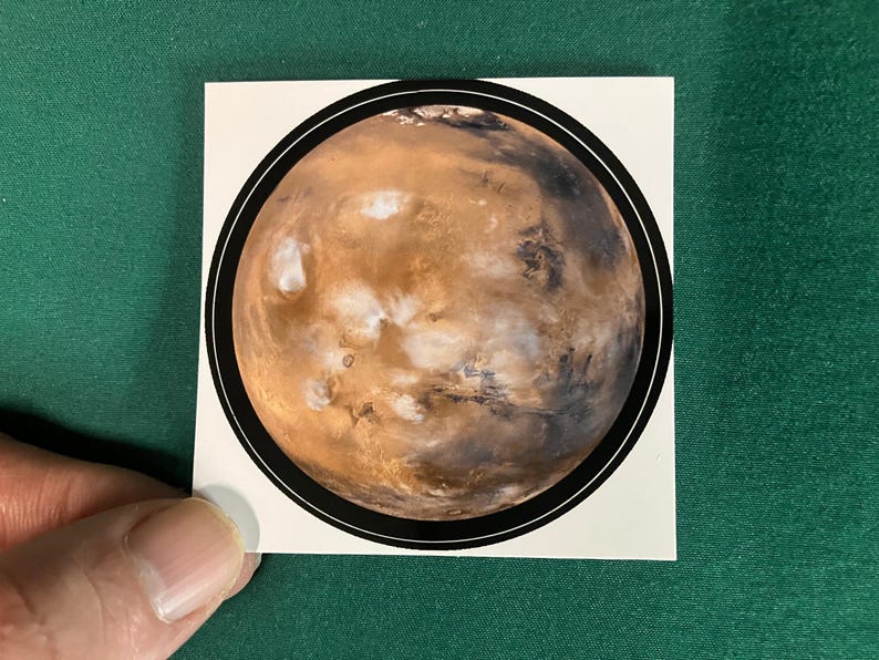 Puede incluir: Una pegatina cuadrada que presenta una ilustraci&oacute;n circular del planeta Marte. El planeta est&aacute; representado en tonos naranja &oacute;xido, marr&oacute;n y blanco, encerrado dentro de un grueso borde circular negro sobre un fondo blanco.