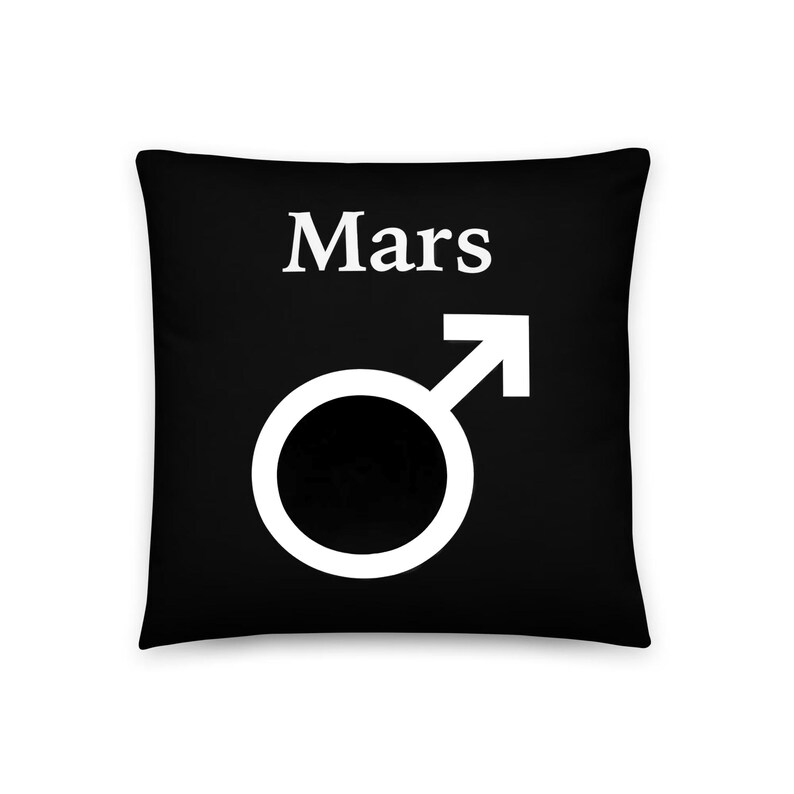 May include: A square black throw pillow featuring the white text Mars centered above a large white Mars planetary symbol, which consists of a circle with an arrow pointing diagonally upward to the right.