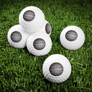 Pode incluir: Um conjunto de cinco bolas de golfe brancas, cada uma com um gr&aacute;fico circular do planeta J&uacute;piter impresso na superf&iacute;cie. As bolas de golfe t&ecirc;m uma textura padr&atilde;o com covinhas e est&atilde;o dispostas em um grupo.