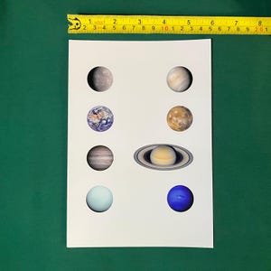 May include: A white sheet featuring eight circular images of planets arranged in two columns, positioned below a yellow measuring tape that shows a width of approximately 8 inches across the top of the paper.
