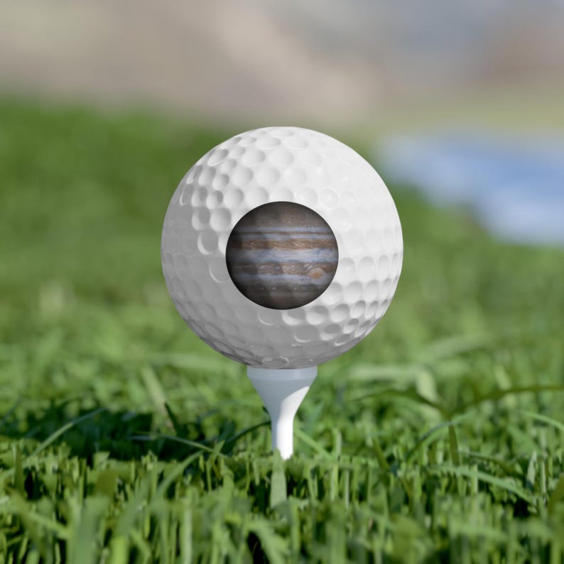 Pode incluir: Uma bola de golfe branca com superf&iacute;cie de covinhas sobre um tee branco. O centro da bola de golfe apresenta uma impress&atilde;o gr&aacute;fica circular que retrata o planeta J&uacute;piter com as suas caracter&iacute;sticas bandas de nuvens castanhas e brancas.